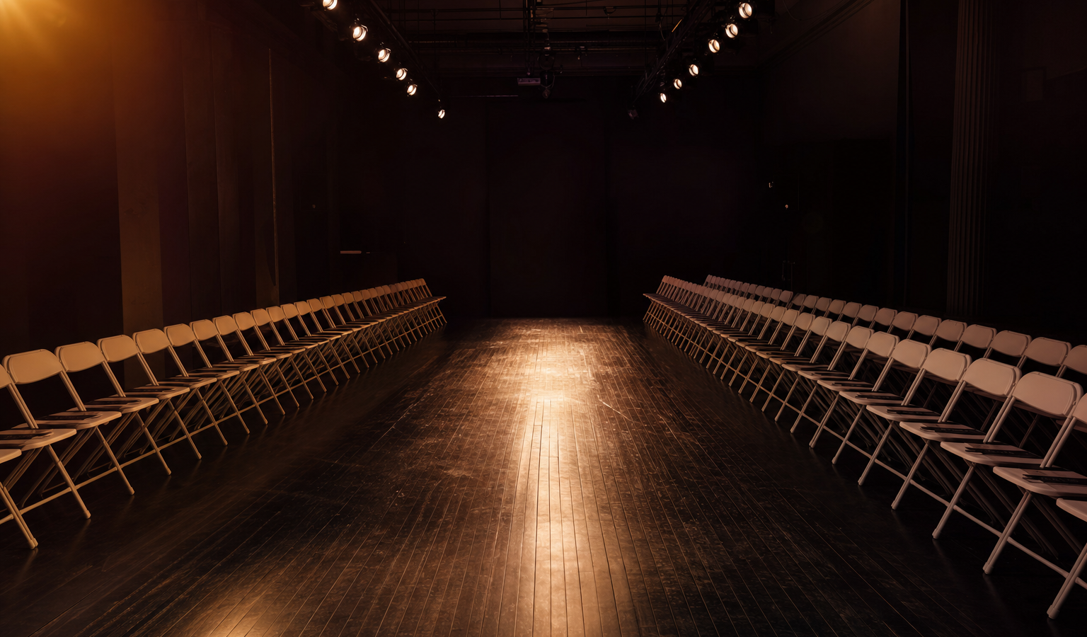 Empty New York Fashion Week runway with warm amber spotlight and folding chairs — the moment before the show