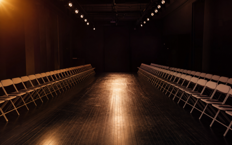 Empty New York Fashion Week runway with warm amber spotlight and folding chairs — the moment before the show