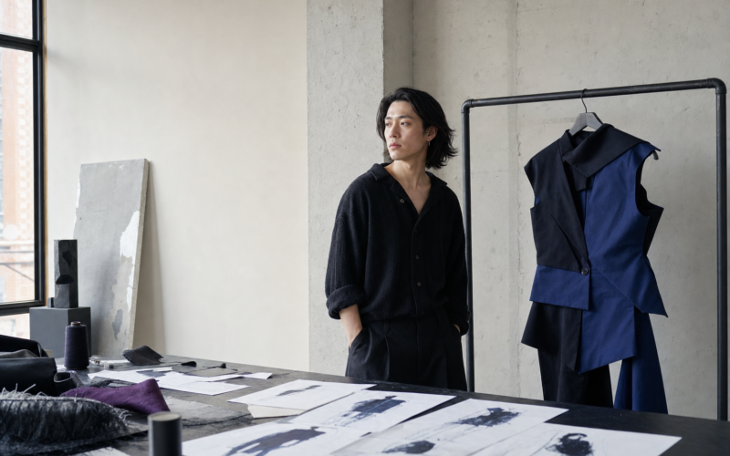 An emerging designer stands beside experimental garments in a minimalist studio.